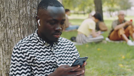 Young-African-Man-in-Earphones-Using-Smartphone-in-Park